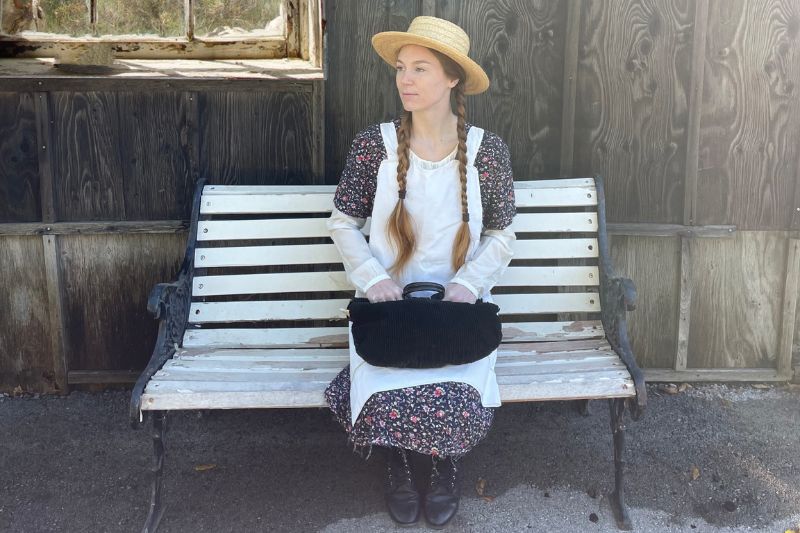 A girl dressed up as Anne of Green Gables wearing a straw hat and sitting on a wooden bench.