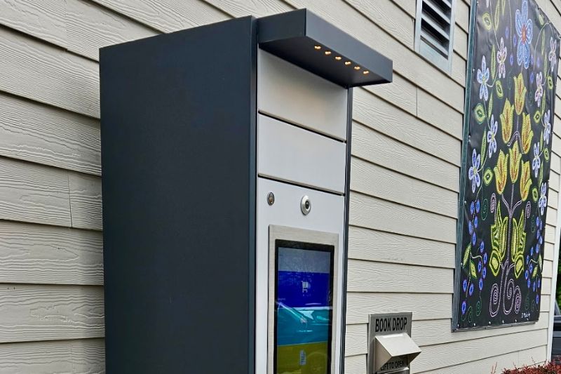 A holds locker unit for picking up library materials standing outside the Maple Acre Branch. a book drop and Indigenous dot art image are in the background.
