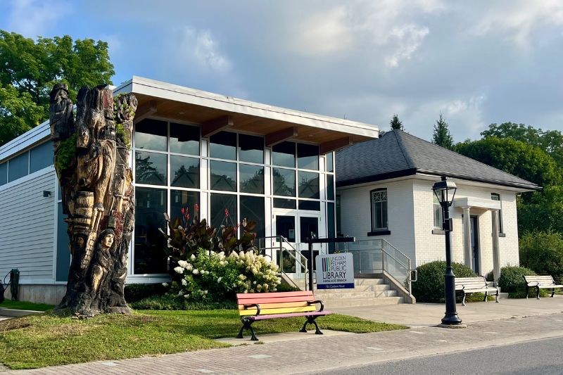 The Maple Acre Branch of the Lincoln Pelham Public Library, which includes a modern glass addition and historic section. A carved tree, rainbow bench, and lamppost are in the foreground.